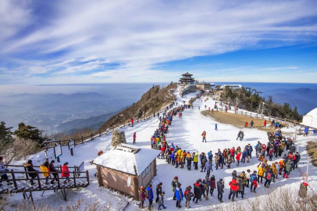 A bustling snowy mountain peak with crowds of tourists in colorful jackets, enjoying the panoramic views and clear blue sky.