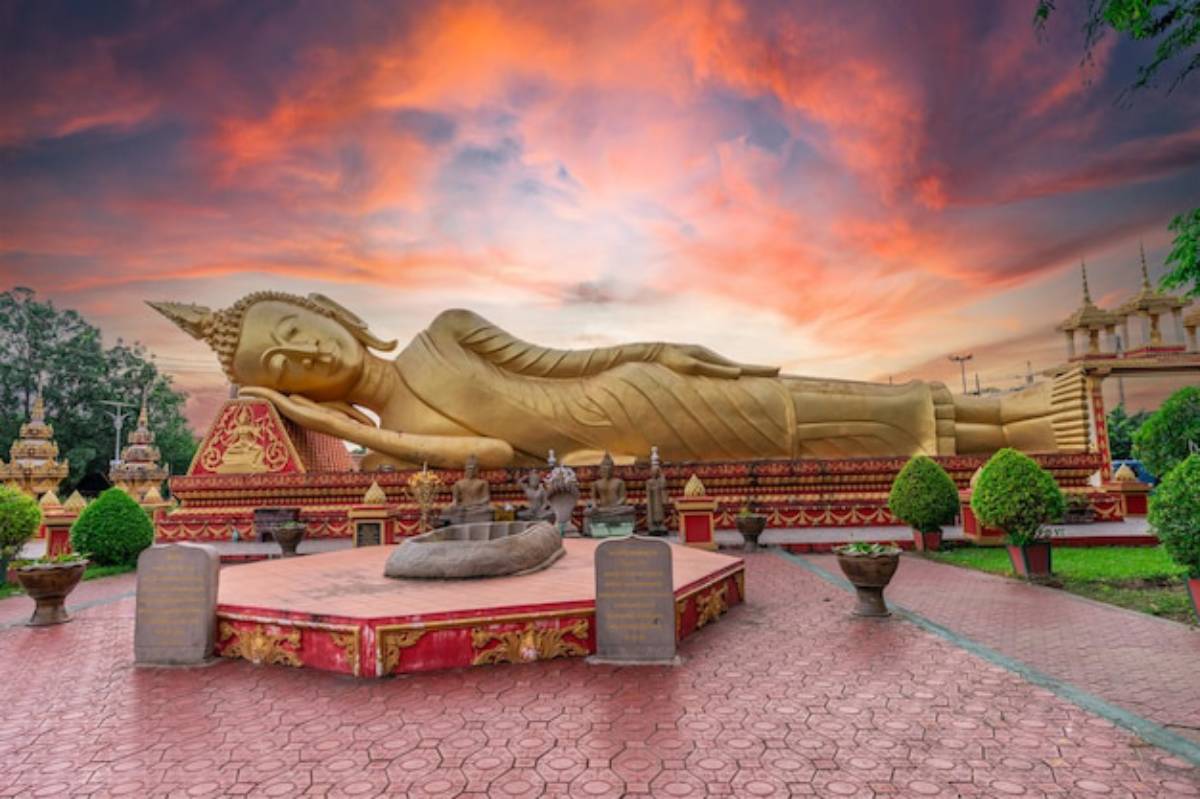 A large golden reclining Buddha statue rests peacefully against a colorful sunset, surrounded by meticulously landscaped gardens.