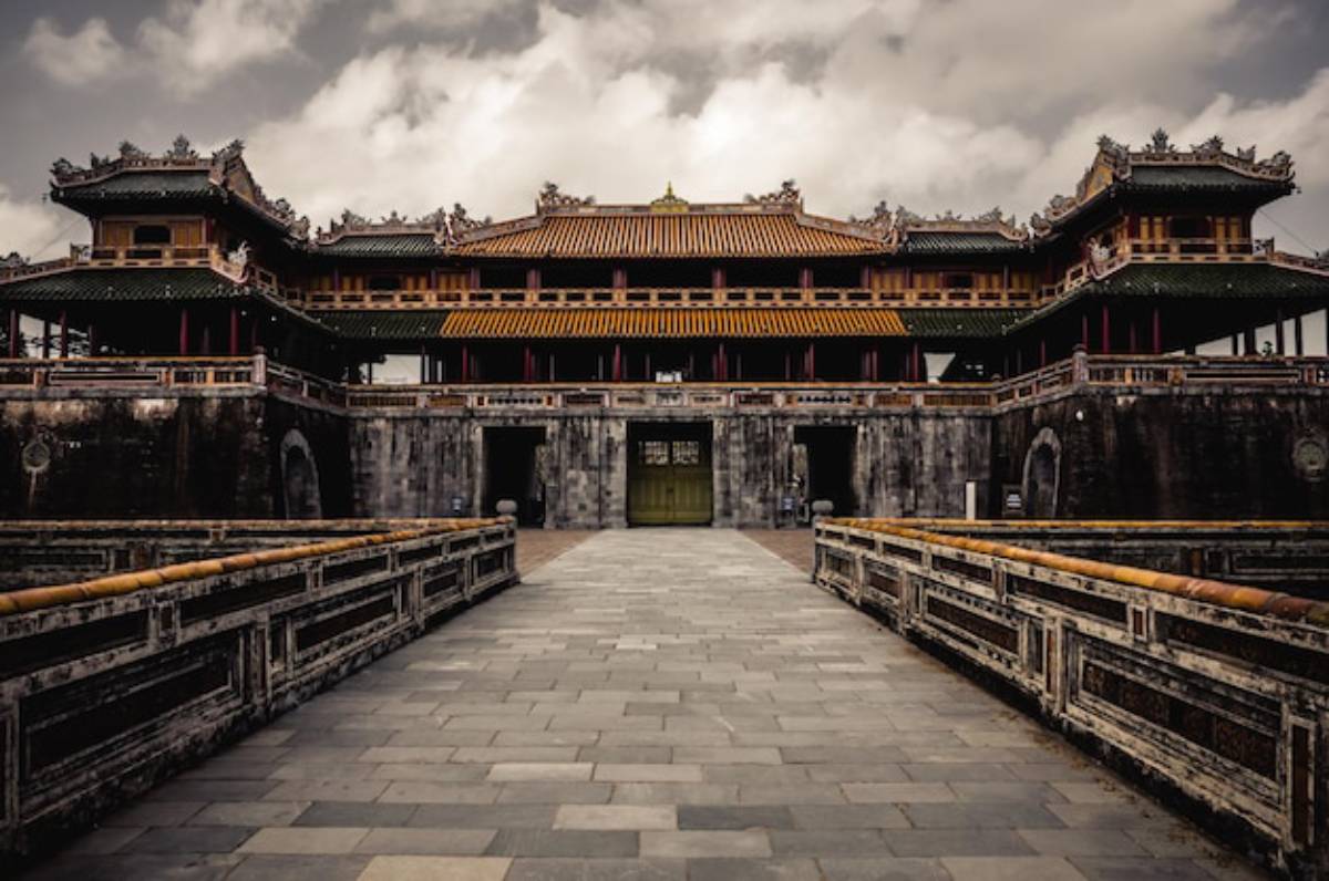 Historic Vietnamese palace with ornate architecture, featuring a stone pathway leading to its grand entrance under a cloudy sky.