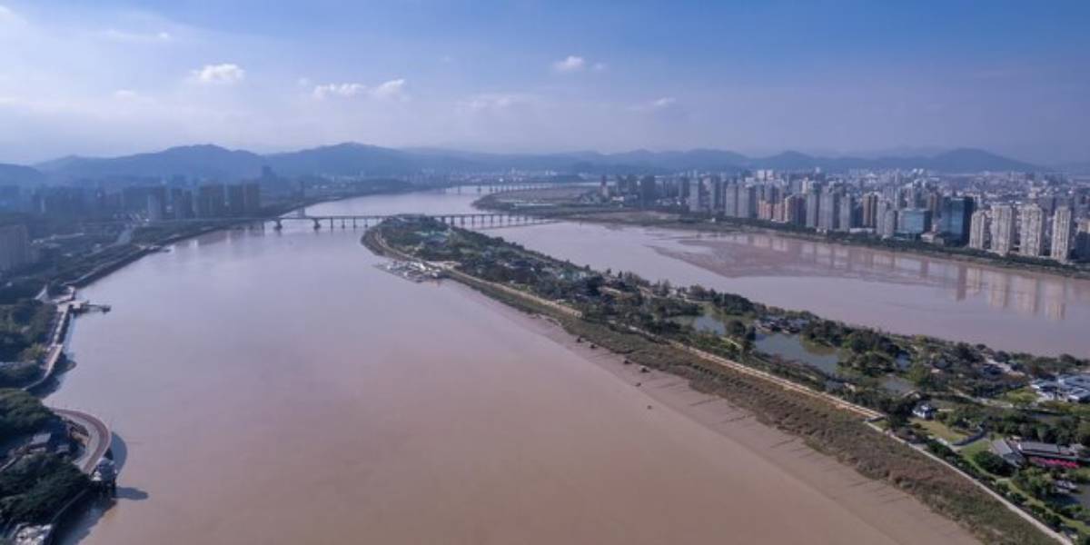 Aerial view of a wide river meandering through a city with bridges, mountains in the background, and urban buildings lining the shore.