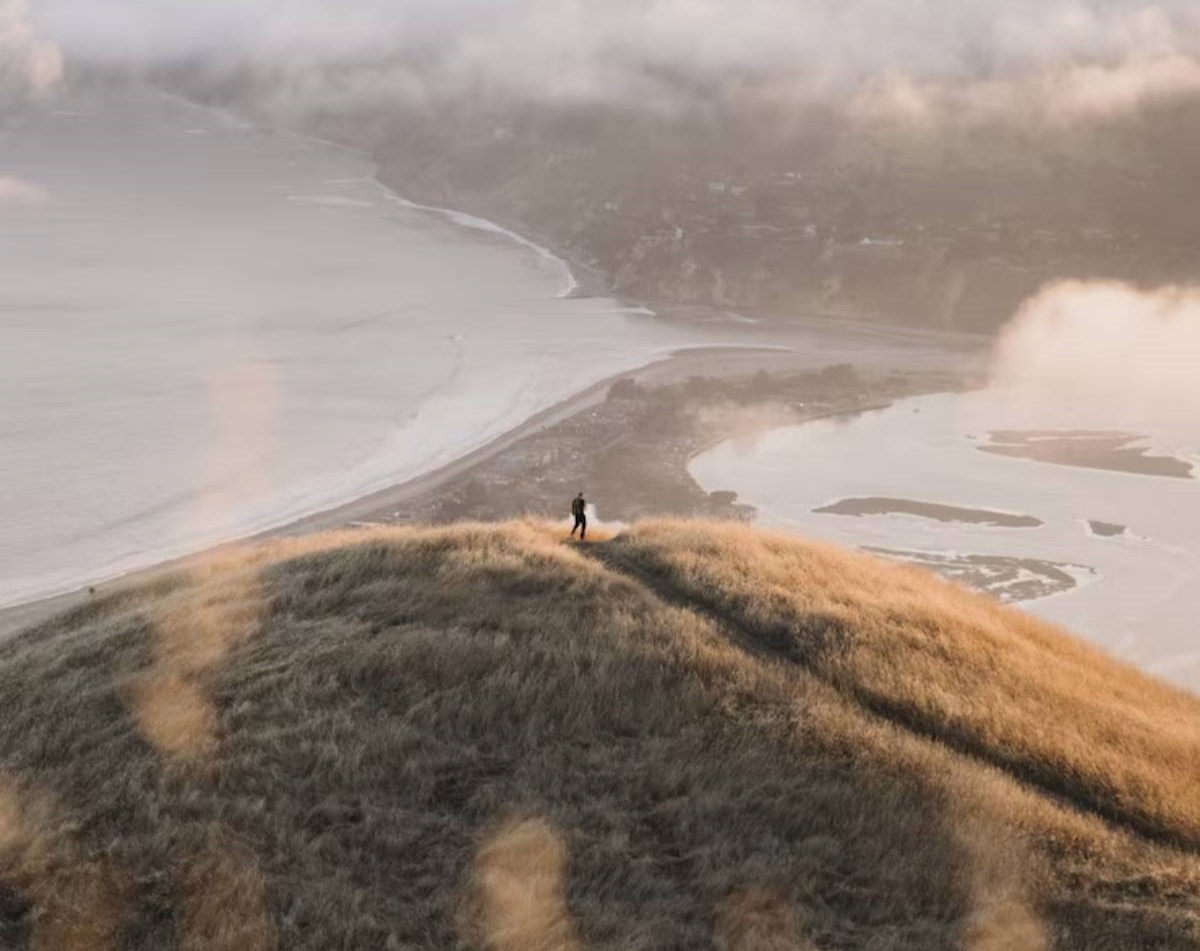 A lone figure stands atop a grassy hill overlooking a misty coastline, with soft clouds blanketing the horizon at sunset.