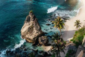 Aerial view of a rocky coastline with turquoise waters, sandy beach, and palm trees under bright sunlight.