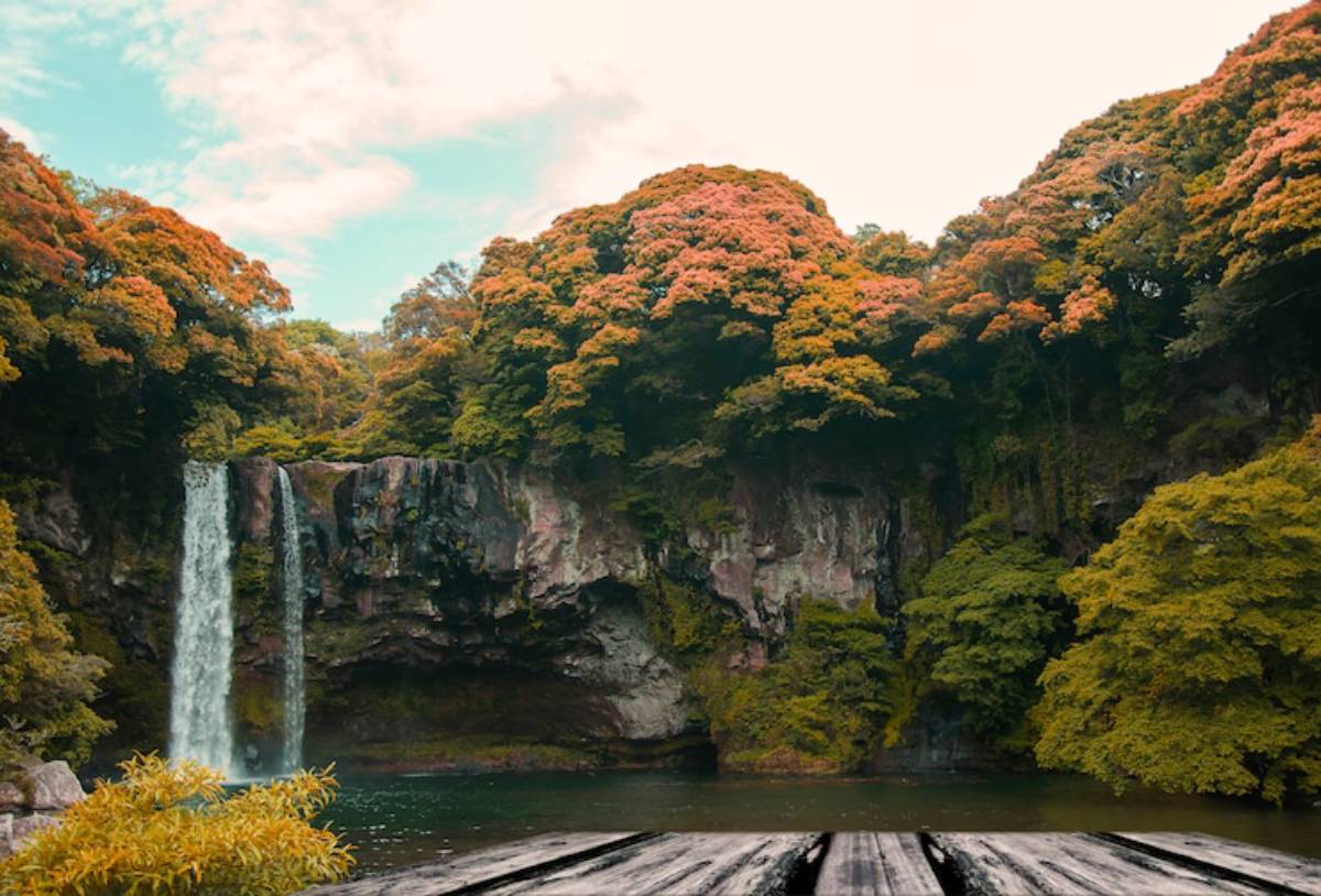 A serene waterfall cascades into a tranquil pool, surrounded by vibrant autumn foliage and rocky cliffs under a soft blue sky.