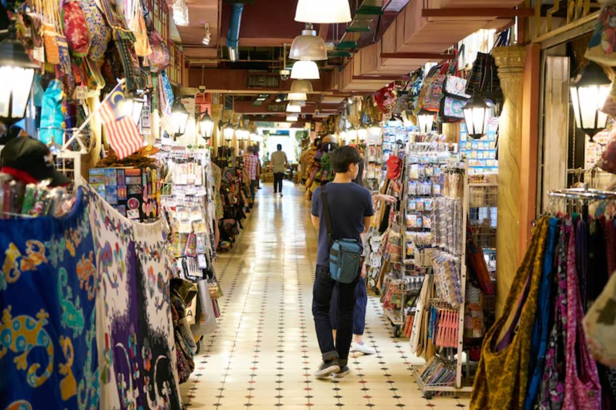 A bustling market corridor filled with colorful souvenirs, fabrics, and shoppers exploring various stalls under warm lighting.