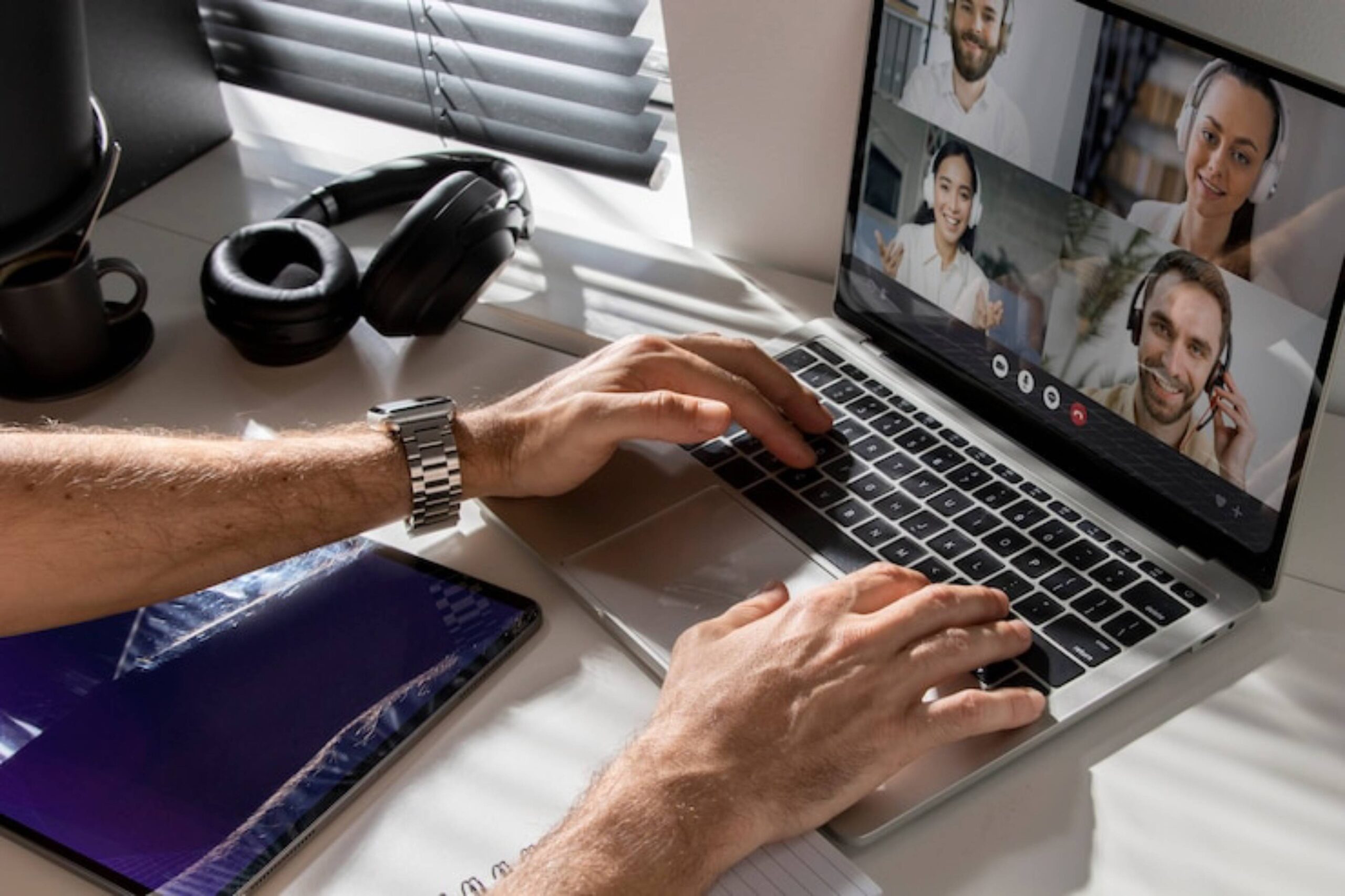 A person typing on a laptop during a video call, with headphones and a notebook visible on a desk illuminated by natural light.