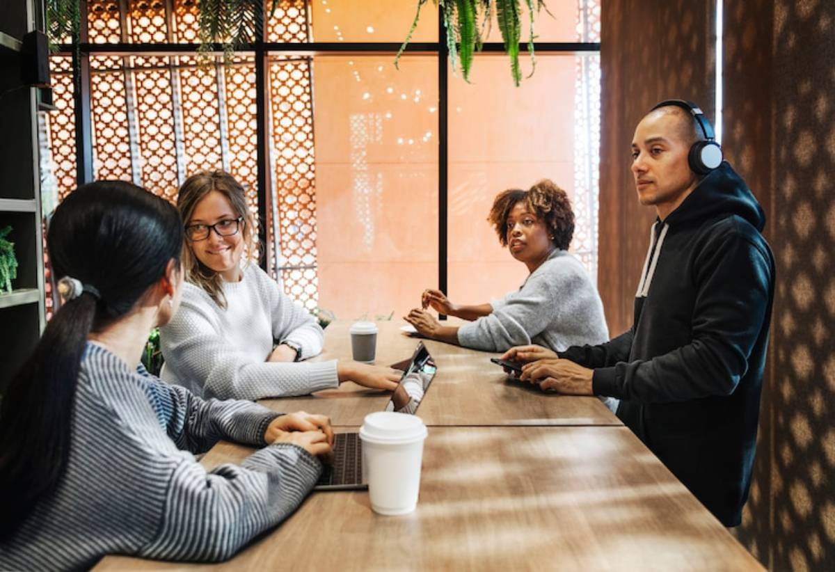 A group of four people engage in conversation and work at a modern wooden table in a cozy cafe with natural light.