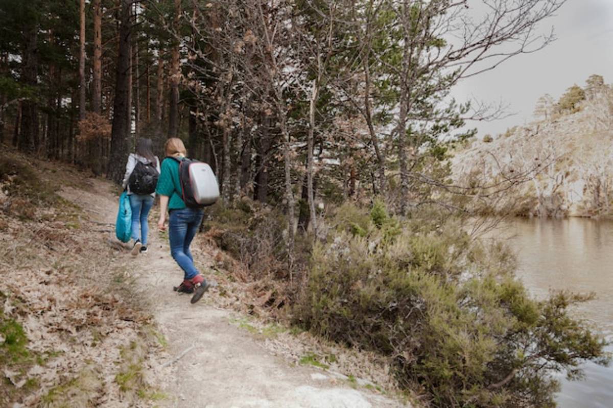 Two hikers with backpacks stroll along a forested trail beside a calm lake, surrounded by tall trees and rocky terrain.