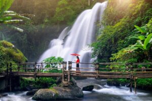 A woman in a red dress and umbrella walks across a bamboo bridge, with a cascading waterfall and lush greenery in the background.