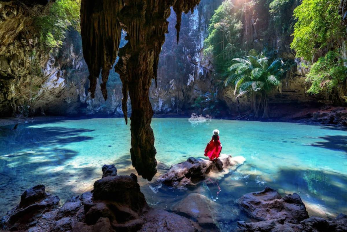 A person in a red dress stands on a rock by a crystal-clear pool in a lush cave, surrounded by greenery and dramatic stalactites.