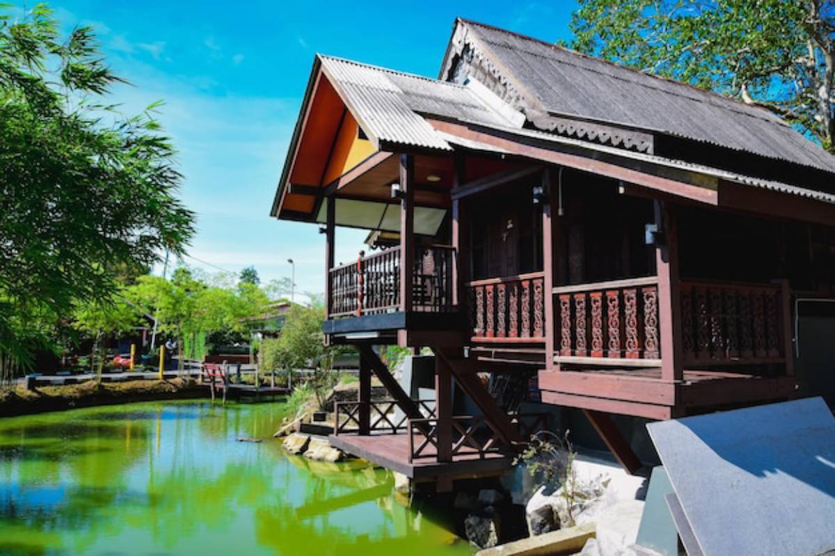 A traditional wooden house on stilts beside a green pond, surrounded by lush trees and a clear blue sky.