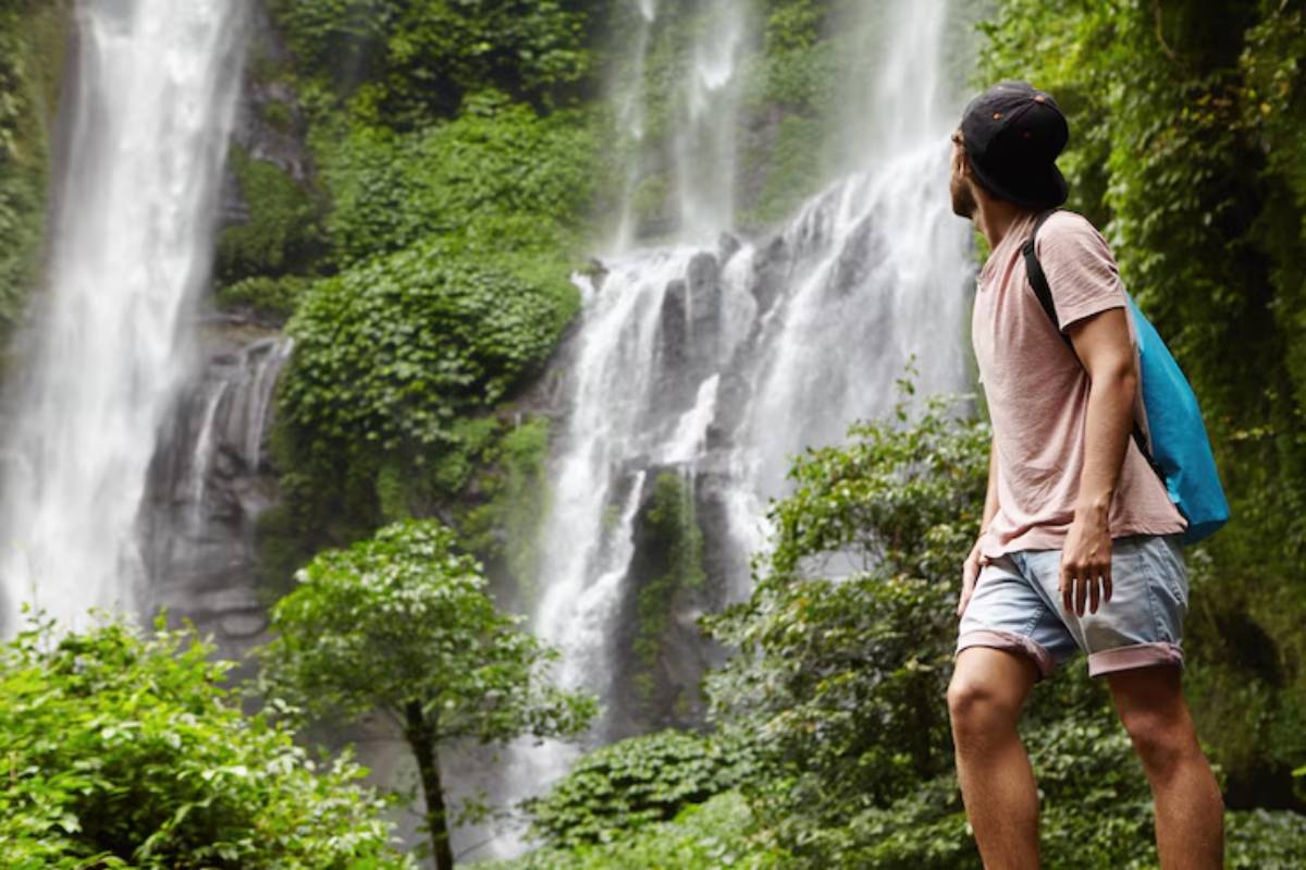A man stands amidst lush greenery, gazing up at a stunning waterfall cascading down rocky cliffs.