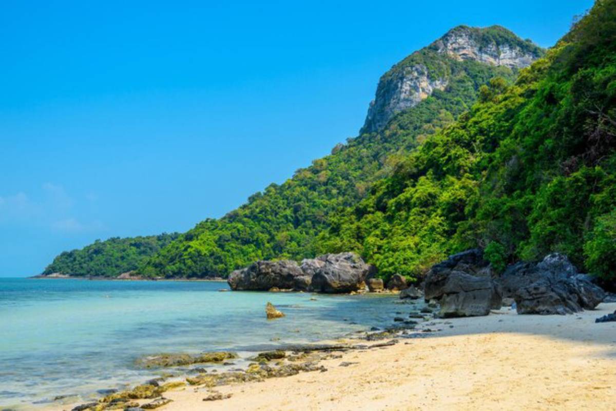 A serene beach with golden sand, clear turquoise waters, and lush green hills rising steeply in the background under a bright blue sky.