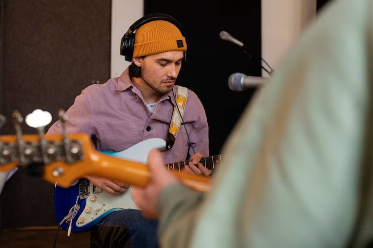 A musician wearing a beanie and headphones plays an electric guitar while another person is in focus, holding a guitar.