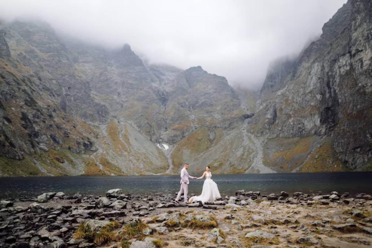 A bride and groom hold hands on rocky shore by a mountain lake, surrounded by fog-shrouded peaks and autumn-colored hills.