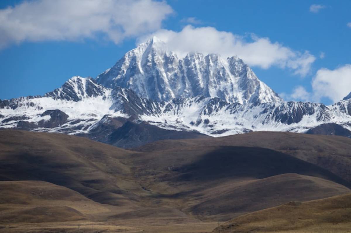 Snow-capped mountain peaks rise majestically against a blue sky, surrounded by rolling brown hills and soft white clouds.