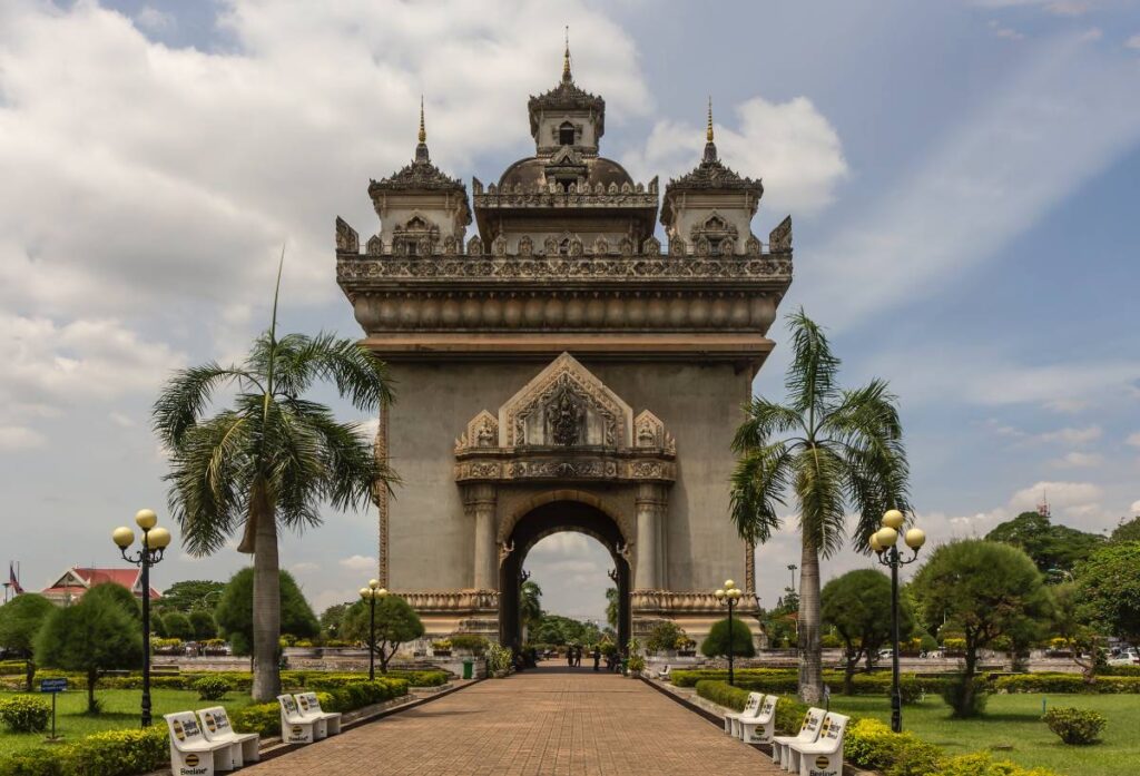 The Patuxai monument in Vientiane, Laos, stands majestically amidst lush gardens under a cloudy sky.