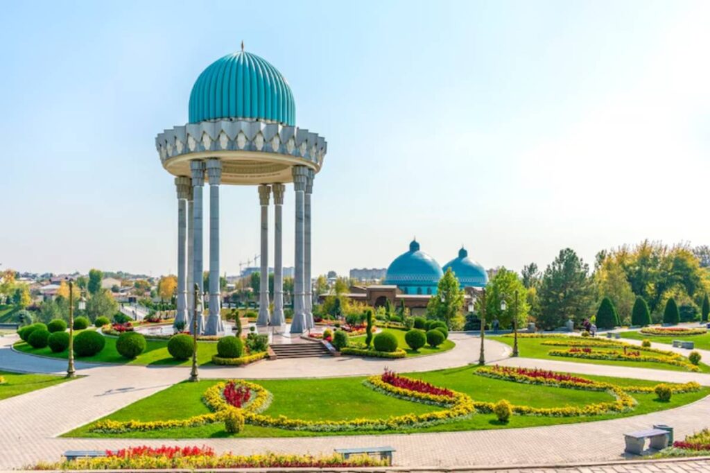 A serene park featuring a grand pavilion with a turquoise dome and manicured gardens adorned with colorful flower beds and pathways.